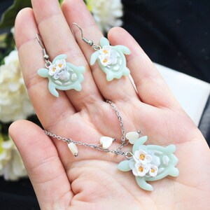 Pode incluir: Um conjunto de joias composto por um colar e um par de brincos pendentes. Cada pe&ccedil;a apresenta um pingente de tartaruga verde-claro adornado com pequenas flores brancas com centros amarelos. O colar inclui uma corrente prateada com cora&ccedil;&otilde;es.