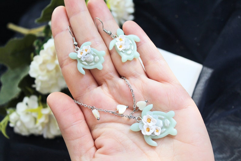 Pode incluir: Um conjunto de joias composto por um colar e um par de brincos pendentes. Cada pe&ccedil;a apresenta um pingente de tartaruga verde-claro adornado com pequenas flores brancas com centros amarelos. O colar inclui uma corrente prateada com cora&ccedil;&otilde;es.