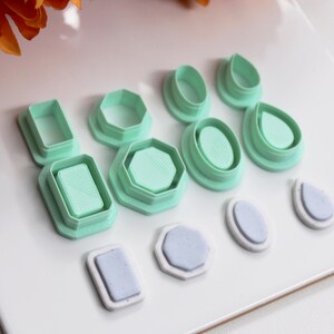 Può includere: Una collezione di taglierini per argilla polimerica in varie forme, tra cui rettangoli, esagoni, ovali e gocce. I taglierini sono verde menta e disposti su una superficie bianca. Alcuni taglierini hanno un inserto viola chiaro. Fiori arancioni sullo sfondo.