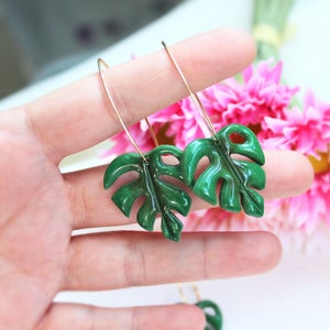 May include: A pair of dangle earrings featuring glossy, dark green monstera leaf charms suspended from thin, gold-toned metal wire hooks. The leaves have a distinct, segmented shape with cutouts, reflecting light on their smooth, polished surface.