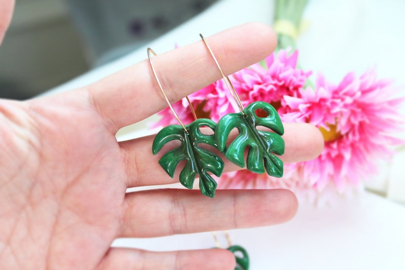 May include: A pair of dangle earrings featuring glossy, dark green monstera leaf charms suspended from thin, gold-toned metal wire hooks. The leaves have a distinct, segmented shape with cutouts, reflecting light on their smooth, polished surface.