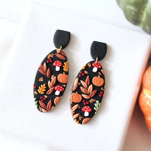 May include: Oval-shaped dangle earrings featuring a black base with a pattern of orange pumpkins, red and white mushrooms, and various autumn-colored leaves. The earrings have a textured black semi-circular top piece connected by a gold-tone ring.
