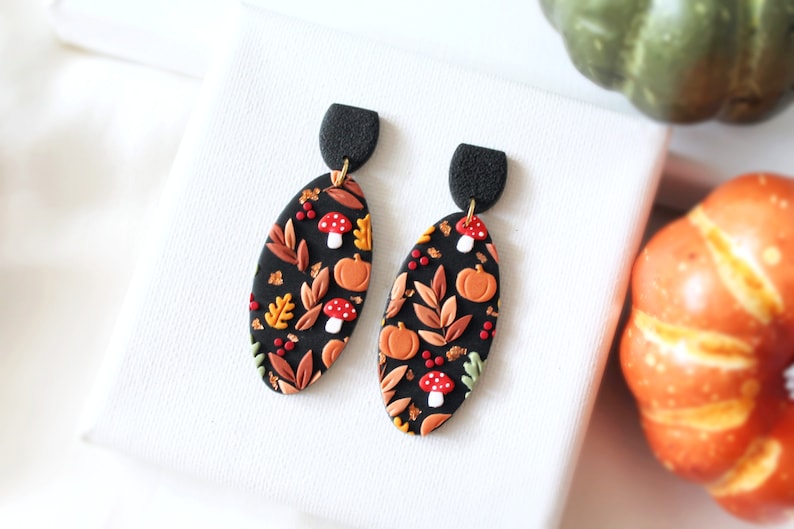 May include: Oval-shaped dangle earrings featuring a black base with a pattern of orange pumpkins, red and white mushrooms, and various autumn-colored leaves. The earrings have a textured black semi-circular top piece connected by a gold-tone ring.
