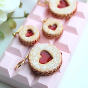May include: Three round cookie charms with a scalloped edge, a heart-shaped cutout in the center, and a red jam filling. Each cookie is dusted with white powder and features a gold-toned metal jump ring attached to the side for hanging.