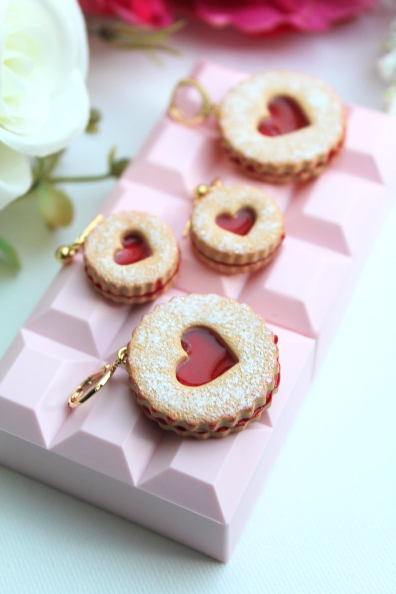 May include: Three round cookie charms with a scalloped edge, a heart-shaped cutout in the center, and a red jam filling. Each cookie is dusted with white powder and features a gold-toned metal jump ring attached to the side for hanging.