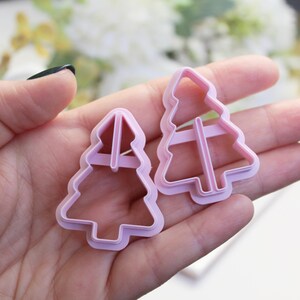 Könnte beinhalten: Zwei hellrosa Kunststoff-Ausstecher in Form eines Weihnachtsbaums. Die Ausstecher werden in einer Hand gehalten, mit einem verschwommenen Hintergrund aus weißen Blumen. Die Ausstecher sind unterschiedlich groß, mit einer einfachen Kontur und inneren Stützen.