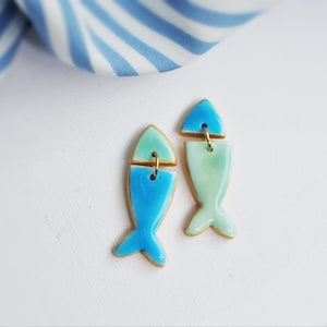 Könnte beinhalten: Ein Paar handgefertigte Ohrringe in Fischform. Die Ohrringe sind blau und grün mit goldfarbenen Akzenten. Die Fischohrringe befinden sich auf einem weißen Hintergrund mit einem blau-weiß gestreiften Stoff im Hintergrund.