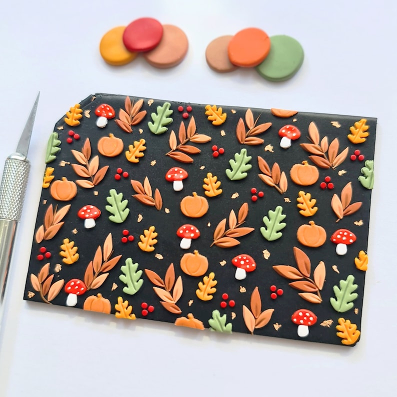May include: A rectangular black polymer clay slab decorated with a repeating pattern of orange pumpkins, red and white mushrooms, green and yellow oak leaves, brown foliage, and small red berries, accented with gold leaf flakes and a craft knife.