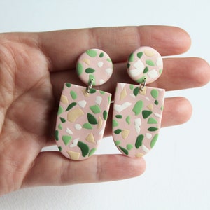 Könnte beinhalten: Ein Paar rosa Terrazzo-Ohrringe mit grünen, weißen und beigefarbenen Akzenten. Die Ohrringe sind aus Polymer Clay gefertigt und haben ein einzigartiges, geometrisches Design.