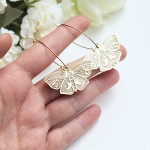 May include: A pair of gold-toned hoop earrings featuring moth-shaped pendants. The moth charms have a metallic gold outline with a light cream-colored enamel inlay, decorated with intricate circular and crescent moon cut-out details.