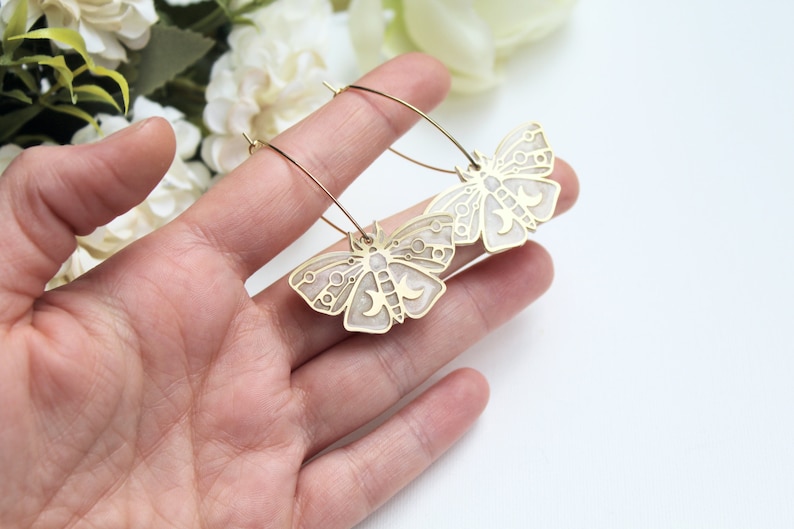 May include: A pair of gold-toned hoop earrings featuring moth-shaped pendants. The moth charms have a metallic gold outline with a light cream-colored enamel inlay, decorated with intricate circular and crescent moon cut-out details.