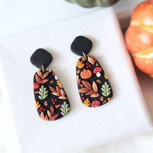 May include: A pair of dangle earrings featuring black, textured diamond-shaped studs connected to elongated, rounded rectangular drops. The drops have a black base decorated with a pattern of orange pumpkins, red mushrooms, and various leaves in shades of green, brown, and yellow.