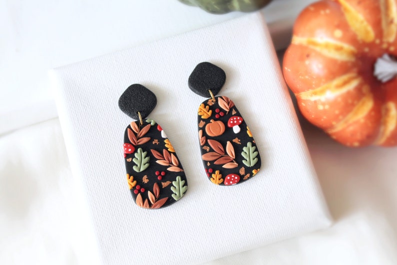 May include: A pair of dangle earrings featuring black, textured diamond-shaped studs connected to elongated, rounded rectangular drops. The drops have a black base decorated with a pattern of orange pumpkins, red mushrooms, and various leaves in shades of green, brown, and yellow.