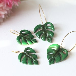 May include: Four hoop earrings featuring glossy, dark green monstera leaf charms. Each earring consists of a thin gold-toned metal hoop attached to a sculpted, segmented leaf pendant with distinct cut-out details and a smooth, reflective finish.