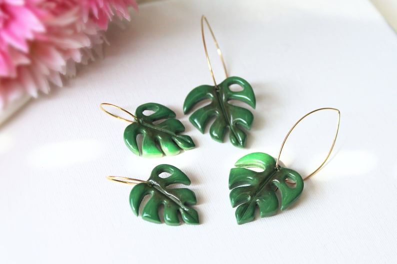 May include: Four hoop earrings featuring glossy, dark green monstera leaf charms. Each earring consists of a thin gold-toned metal hoop attached to a sculpted, segmented leaf pendant with distinct cut-out details and a smooth, reflective finish.