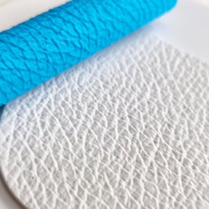 Può includere: Un mattarello cilindrico blu brillante con un motivo a griglia a forma di diamante impresso sulla superficie, posizionato accanto a un pezzo di materiale circolare bianco che mostra un'impronta testurizzata dello stesso motivo.