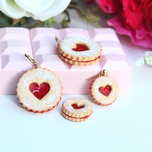 May include: A collection of four heart-shaped sandwich cookie charms featuring a light tan biscuit exterior dusted with white powder and a bright red translucent jam center. The charms vary in size and include metal hardware for attachment.