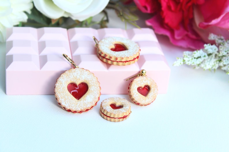 May include: A collection of four heart-shaped sandwich cookie charms featuring a light tan biscuit exterior dusted with white powder and a bright red translucent jam center. The charms vary in size and include metal hardware for attachment.