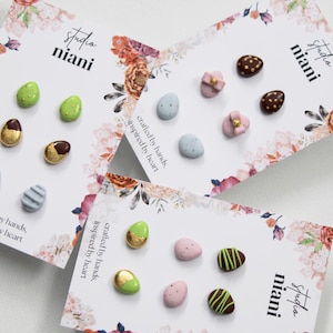 May include: A selection of Easter egg ornaments in shades of green, blue, pink, and brown, some with gold detailing. The cards feature floral designs and the text "studio niani" and "crafted by hands, inspired by heart."