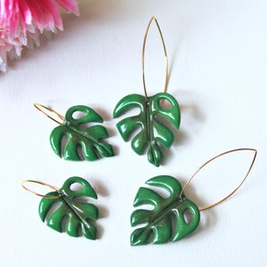 May include: Four green monstera leaf-shaped charms attached to thin gold-toned metal earring hooks. The leaves have a glossy finish with cut-out details, arranged in two pairs of varying sizes on a plain white surface.