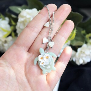Pode incluir: Um colar de corrente de metal prateado com um pingente de tartaruga marinha verde-claro adornado com tr&ecirc;s pequenas flores brancas com centros amarelos. A corrente inclui dois pequenos pingentes brancos em forma de cora&ccedil;&atilde;o.