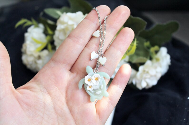 Pode incluir: Um colar de corrente de metal prateado com um pingente de tartaruga marinha verde-claro adornado com tr&ecirc;s pequenas flores brancas com centros amarelos. A corrente inclui dois pequenos pingentes brancos em forma de cora&ccedil;&atilde;o.