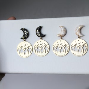 May include: A pair of gold earrings with a crescent moon charm and a cut-out design of a mountain range with stars and a crescent moon.