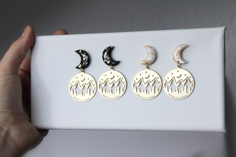 May include: A pair of gold earrings with a crescent moon charm and a cut-out design of a mountain range with stars and a crescent moon.