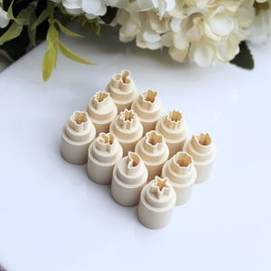 Könnte beinhalten: Ein Set mit 12 kleinen, weißen Plätzchenausstechern mit verschiedenen Blumen- und Blattmotiven. Die Ausstecher sind übereinander gestapelt.