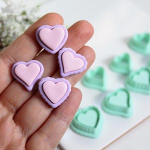 Op de afbeelding: Een verzameling hartvormige polymeerklei versieringen in roze en lavendel. Er zijn ook verschillende hartvormige uitsteekvormen in lichtgroen te zien. Deze knutselbenodigdheden zijn ideaal voor doe-het-zelf projecten.