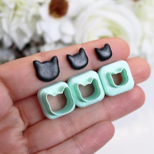 Può includere: Tre piccoli pezzi di argilla nera a forma di gatto disposti in fila sopra tre stampini quadrati color verde menta chiaro con ritagli a forma di gatto al centro. Gli oggetti sono abbastanza piccoli da essere tenuti nel palmo di una mano.