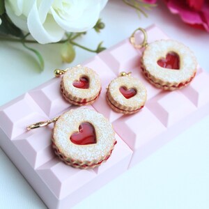 May include: Four round cookie-shaped charms featuring a textured beige surface with a central heart-shaped cutout revealing a translucent red filling. Each charm has a small gold-toned metal hoop attached to the top edge for hanging purposes.