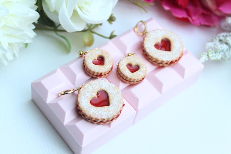 May include: Four round cookie-shaped charms featuring a textured beige surface with a central heart-shaped cutout revealing a translucent red filling. Each charm has a small gold-toned metal hoop attached to the top edge for hanging purposes.
