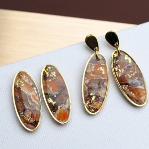 Könnte beinhalten: Ein Paar goldene ovale Ohrringe mit einem braunen und goldenen Marmormuster. Die Ohrringe haben einen goldenen Hakenverschluss.