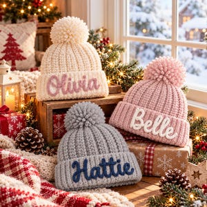 Op de afbeelding: Drie gebreide mutsen met pompons, elk voorzien van een naam in reliëf. De crèmekleurige muts toont Olivia, de roze muts Belle en de grijze muts Hattie. Elke muts heeft een geribbelde textuur en een omgeslagen boord.