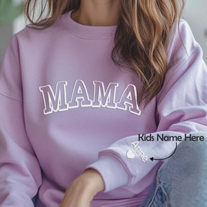 May include: A lavender sweatshirt with the word "MAMA" in white outlined letters on the front. The left sleeve has a small heart with the word "Anne" embroidered below it. The text "Kids Name Here" is written above the heart.