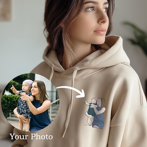 Może przedstawiać: Beżowa bluza z kapturem z haftowanym wzorem matki i dziecka. Bluza z kapturem ma kaptur ze sznurkiem i kieszeń typu kangur. Słowa "Your Photo" są wydrukowane w lewym dolnym rogu obrazu. Nazwa marki "Lamoriea" jest wydrukowana w prawym górnym rogu.