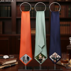 May include: Three neckties in orange, light green, and navy blue, each featuring a square photo print at the bottom tip. The ties are suspended from silver metal rings on individual stands. The brand name LAMORIEA is visible in the top left corner.