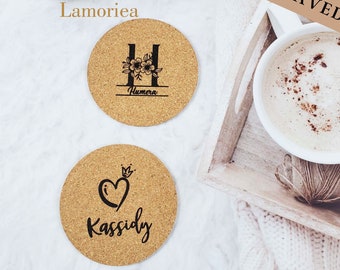 May include: Two round cork coasters with laser-engraved designs. One features the letter "H" with floral accents and the name "Humera". The other has a heart with a crown and the name "Kassidy". The text "PERMANENT LASER ENGRAVED" is also visible.