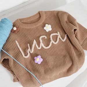 May include: A brown long-sleeve knit jumper featuring the name "Luca" in cream-coloured yarn lettering across the chest. The jumper is decorated with three small, crocheted flower appliqu&eacute;s in white, brown, and purple, and sits next to a ball of blue yarn.