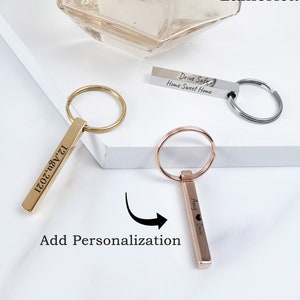 Peut inclure: Deux porte-clés dorés avec des barres gravées personnalisées. Une barre porte l'inscription "12, Agu, 2021" et l'autre "Amy Dave" avec un symbole de cœur. Les porte-clés sont sur une surface blanche avec le texte "Ajouter une personnalisation" en dessous.