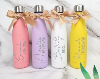 Könnte beinhalten: Vier personalisierte Edelstahl-Wasserflaschen mit goldenen Schleifen. Die Flaschen sind rosa, lila, weiß und gelb. Die Flaschen sind mit den Namen "Sarah Bridesmaid", "Amelia Bridesmaid", "Brittany Bride" und "Hannah Bridesmaid" beschriftet.