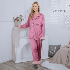 May include: A pink satin pyjama set with white trim, featuring a button-down top and matching trousers. The top has embroidered initials and feather trim on the cuffs. The pyjama set is from Lamoriea.