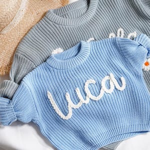 May include: A light blue knit jumper featuring the name Luca in white cursive yarn lettering, layered over a grey knit jumper with a white daisy flower detail, accompanied by a tan straw hat with a frayed edge and a white ribbon trim.