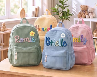 May include: Four corduroy backpacks in sage green, yellow, light blue, and pink. Each bag features a front zipped pocket, side pockets, and a name embroidered in colourful, raised script lettering. A small fabric flower is attached to the front.
