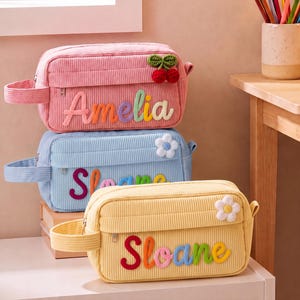 May include: Three rectangular corduroy pencil cases in pink, blue, and yellow. The pink case features a cherry applique and the name Amelia. The blue and yellow cases feature flower appliques and the name Sloane. Each has a side handle and zipper.