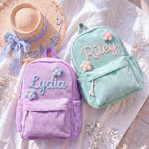 May include: A purple corduroy backpack with the name Lydia and two blue flower patches, and a mint green corduroy backpack with the name Riley and two pink flower patches, both featuring front zippered pockets and top carry handles.