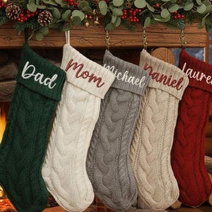 Könnte beinhalten: Eine gemütliche Weihnachtsszene mit fünf Zopfstrick-Strümpfen, die an einem Holzkamin hängen. Jeder Strumpf hat eine andere Farbe: Grün, Weiß, Grau, Beige und Rot, mit eingestickten Namen. Im Hintergrund ein Kamin mit brennendem Feuer.