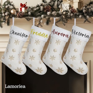 Pode incluir: Quatro meias de Natal brancas com padrões de flocos de neve dourados e nomes personalizados: Jennifer, Freddie, Aurora e Victoria.
