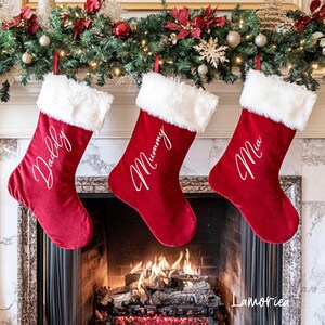 Op de afbeelding: Drie rode fluwelen kerstsokken met witte nep bont trim en witte letters. De sokken hangen aan een schoorsteenmantel met een brandend vuur in de open haard. De sokken zijn gepersonaliseerd met de namen "Daddy", "Mommy" en "Mia".